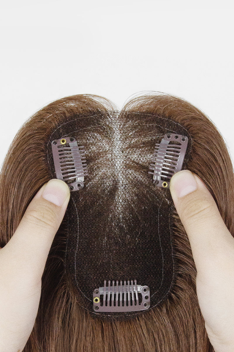 A close-up of a brown hairpiece or wig with three dark mesh clip-in sections held in place by two hands against a plain white background.