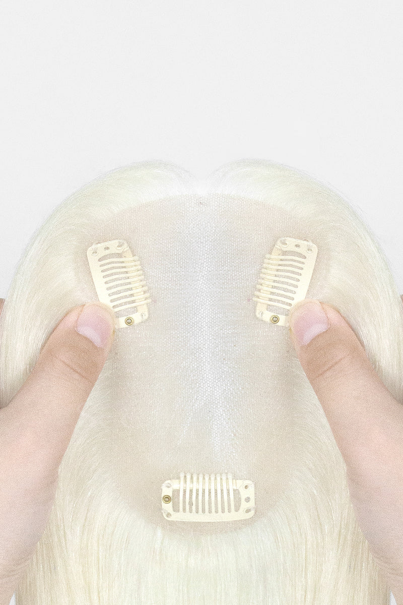 A close-up of a blonde wig with three cream-colored hair clip-in combs attached, held by two hands against a plain white background.