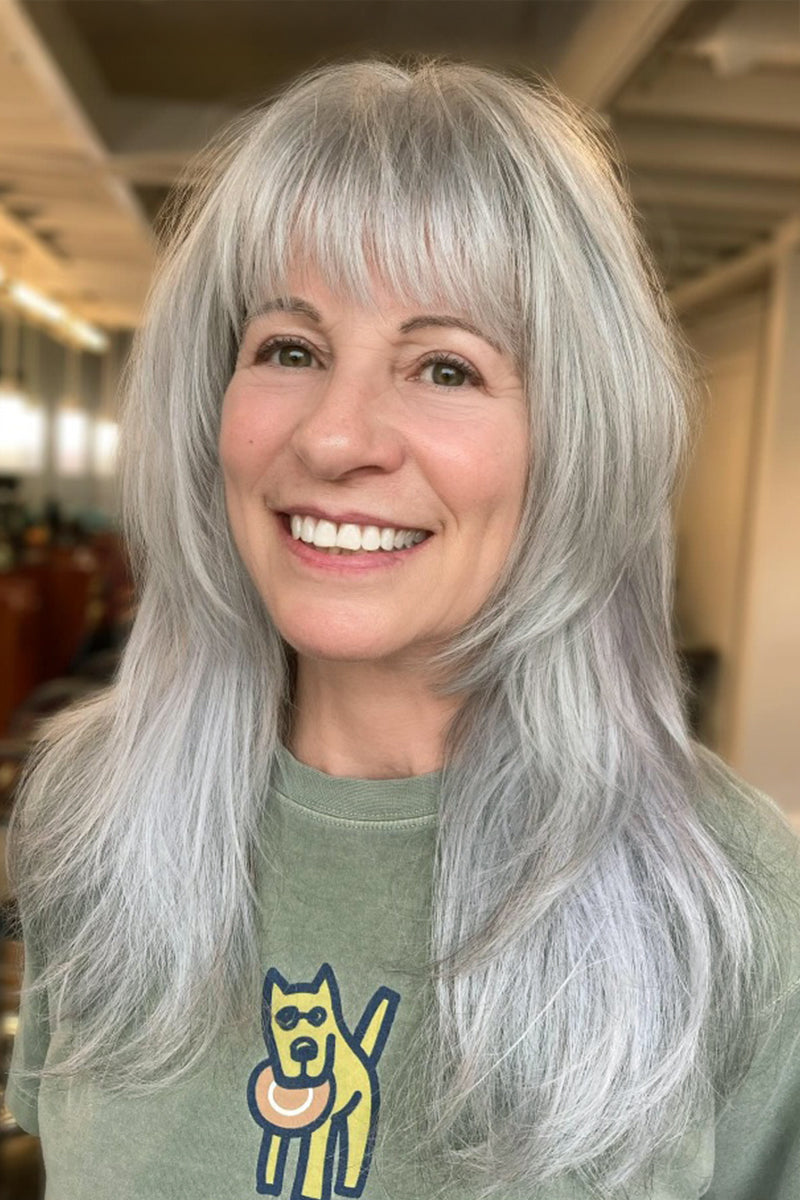 A smiling woman with long, wavy silver-gray hair and bangs, wearing a sage green t-shirt with a yellow and blue dog graphic, photographed in an indoor setting with a blurred background.