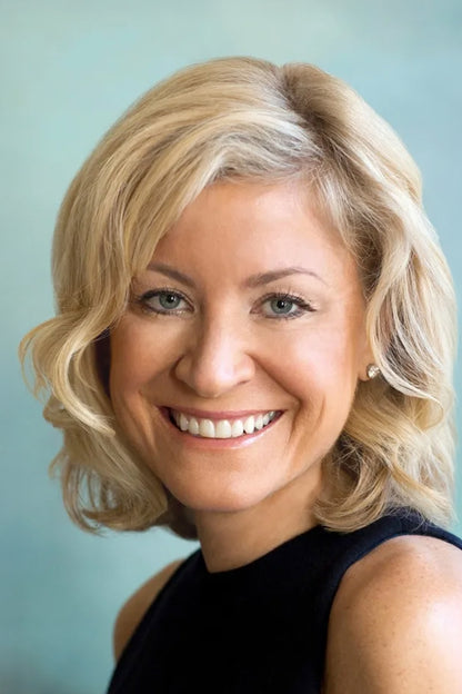 A smiling blonde woman with blue eyes wearing a black sleeveless top, photographed against a soft light blue background.