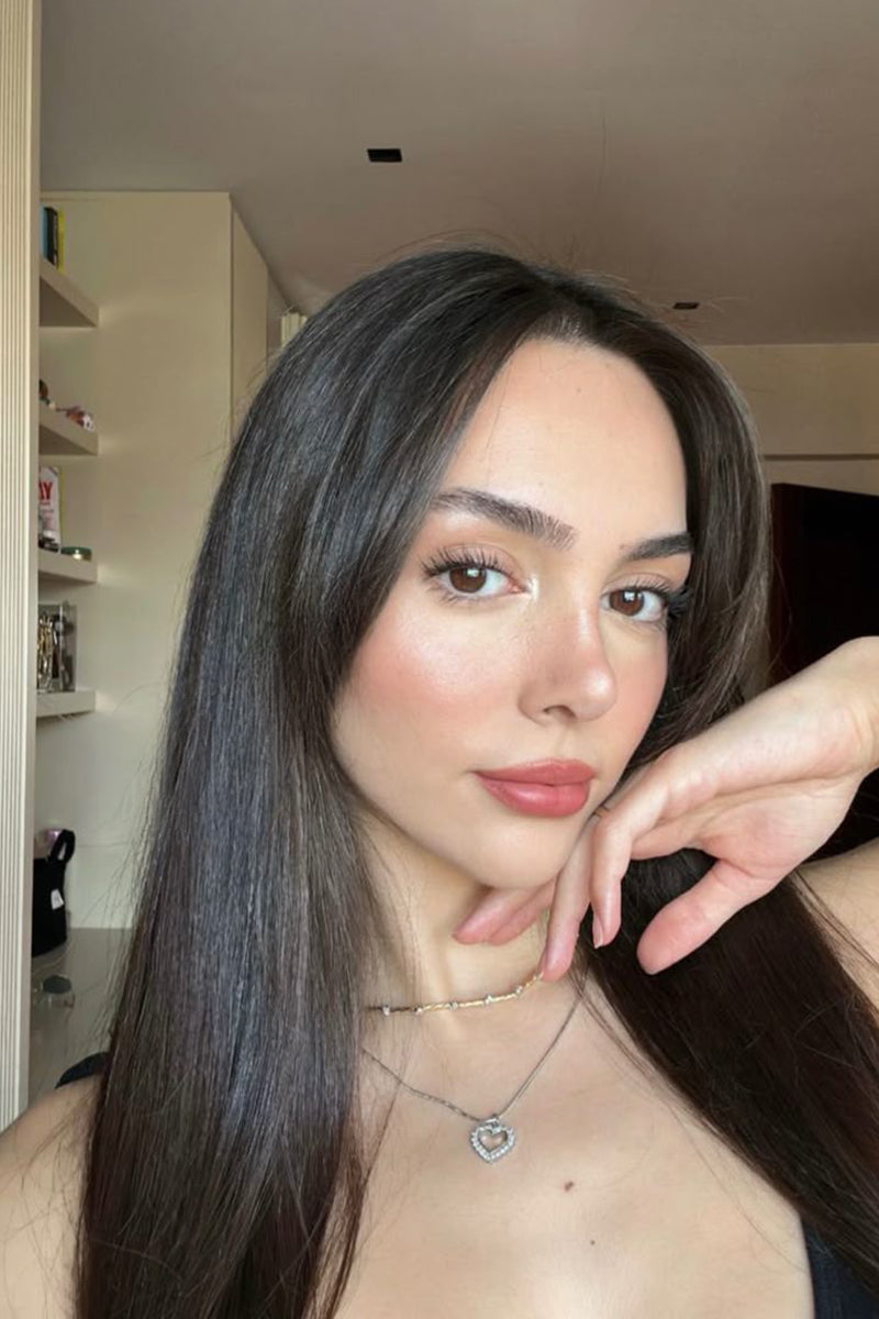 A close-up portrait of a woman with long, straight dark brown hair wearing a delicate silver necklace with a heart-shaped pendant. She is posing indoors with her hand resting near her chin.