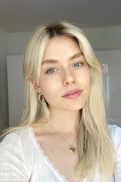 A close-up portrait of a young woman with long blonde hair, wearing gold hoop earrings and a delicate gold heart necklace, in a brightly lit indoor setting.