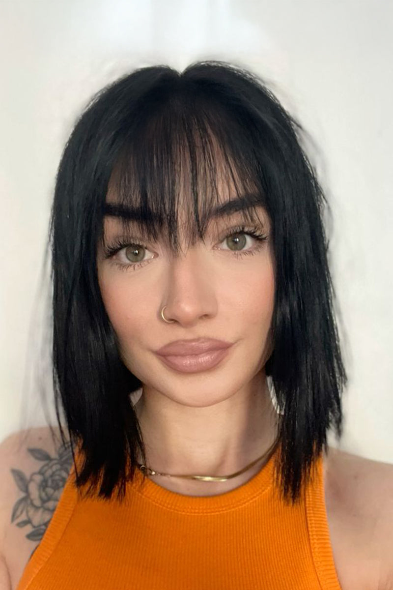 A close-up portrait of a woman with a short, straight black bob haircut and bangs, wearing an orange ribbed tank top against a plain light background.