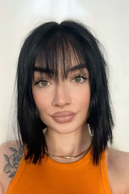 A close-up portrait of a woman with a short, straight black bob haircut and bangs, wearing an orange ribbed tank top against a plain light background.