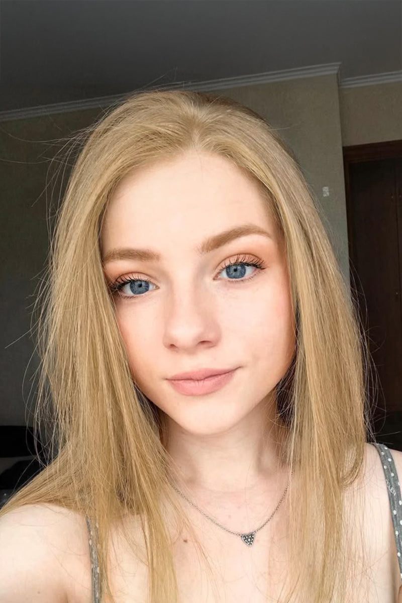 A close-up selfie of a young woman with long blonde hair, blue eyes, and a neutral expression, wearing a delicate silver necklace with a small heart pendant.
