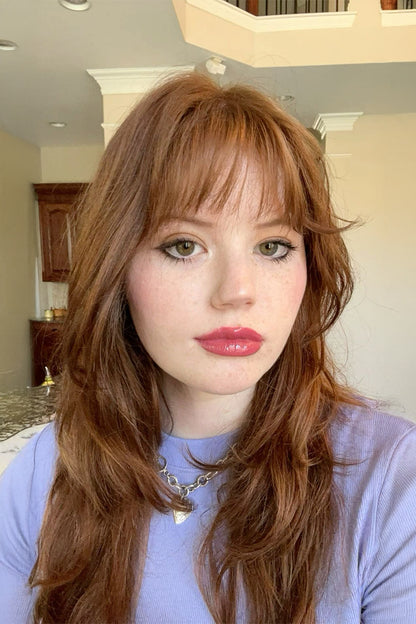 A woman with long, wavy auburn-brown hair and bangs, wearing a lavender top and a silver chain necklace, photographed in a selfie-style close-up inside a home.