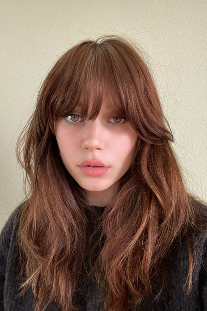 A close-up portrait of a woman with shoulder-length, wavy auburn-brown hair and straight bangs, wearing a dark gray knit top against a plain beige wall.