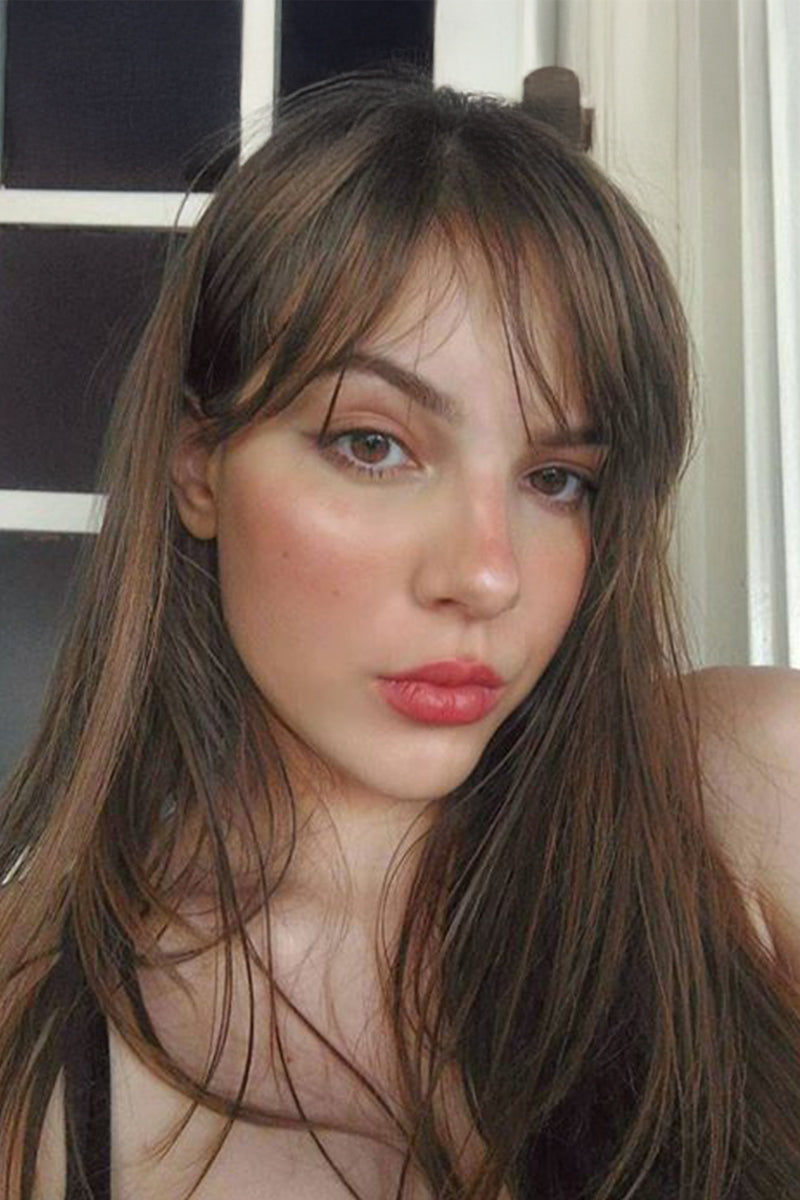 A close-up portrait of a woman with long, straight brown hair and bangs, wearing pink lipstick, set against an indoor background with a white-framed window.