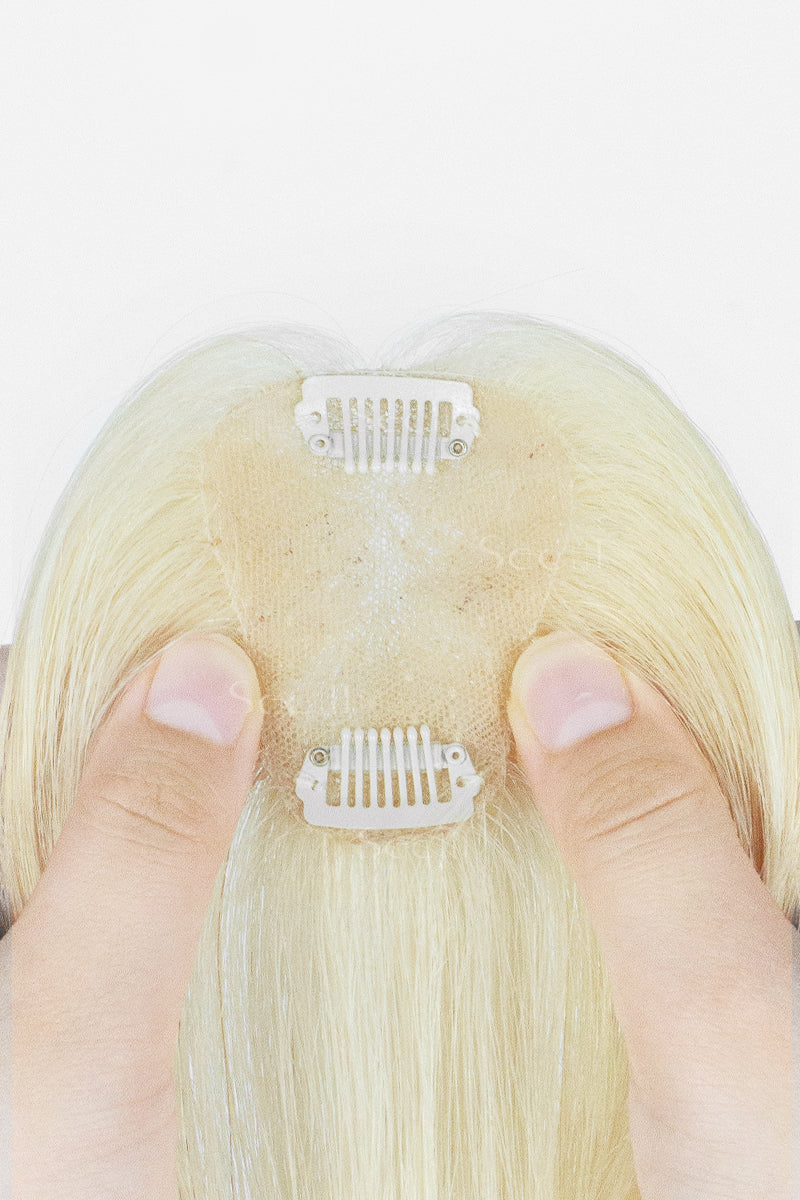 Close-up of the back of a blonde wig or hairpiece with two white clip-in sections attached to a beige lace-like inner cap, held by two fingers against a white background.