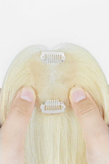 Close-up of the back of a blonde wig or hairpiece with two white clip-in sections attached to a beige lace-like inner cap, held by two fingers against a white background.