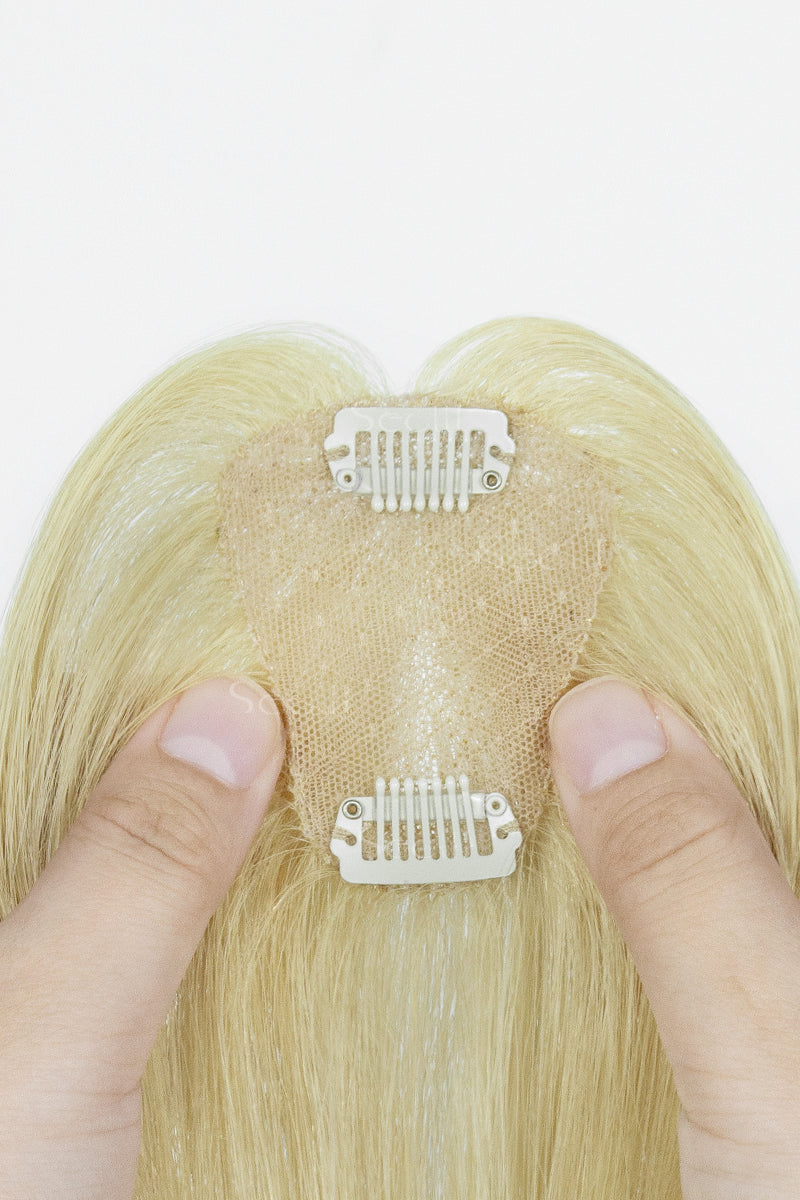 A close-up of a blonde hairpiece with a beige adhesive mesh crown and two white clip attachments, held by two fingers against a plain white background.