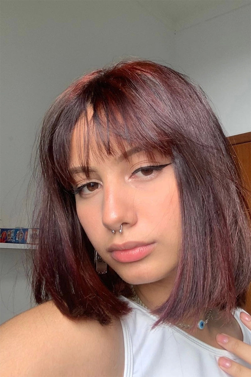 A close-up selfie of a person with shoulder-length, straight, dark auburn hair with bangs, wearing a white top and a nose ring.