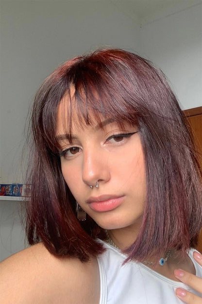 A close-up selfie of a person with shoulder-length, straight, dark auburn hair with bangs, wearing a white top and a nose ring.