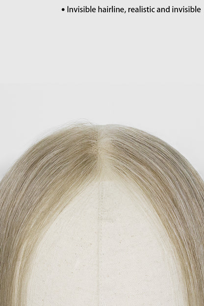 Close-up of a wig or hairpiece on a mannequin head, showing a center part with light ash blonde to beige hair against a plain light background. Small text at the top reads &