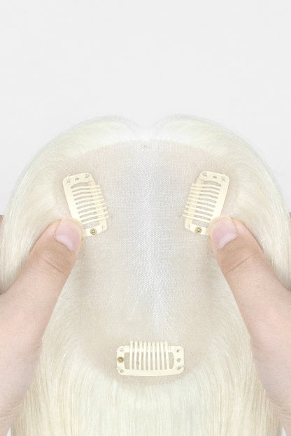 A close-up of a blonde wig with three cream-colored hair clip-in combs attached, held by two hands against a plain white background.