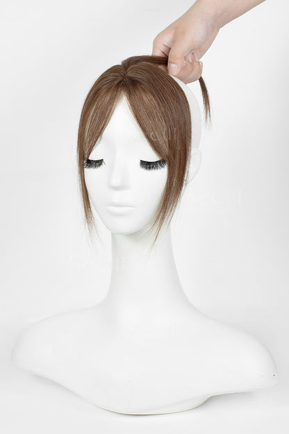 A brown topper with straight bangs displayed on a white mannequin head, with a hand lifting the hair at the top against a plain white background.