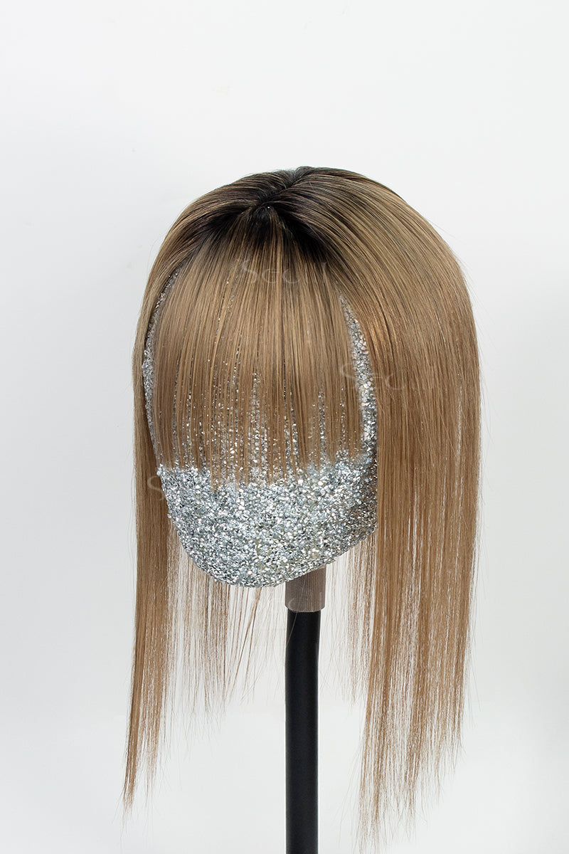 A mannequin head displaying a handmade human hair topper with bangs, featuring a natural black root and medium ash brown color.