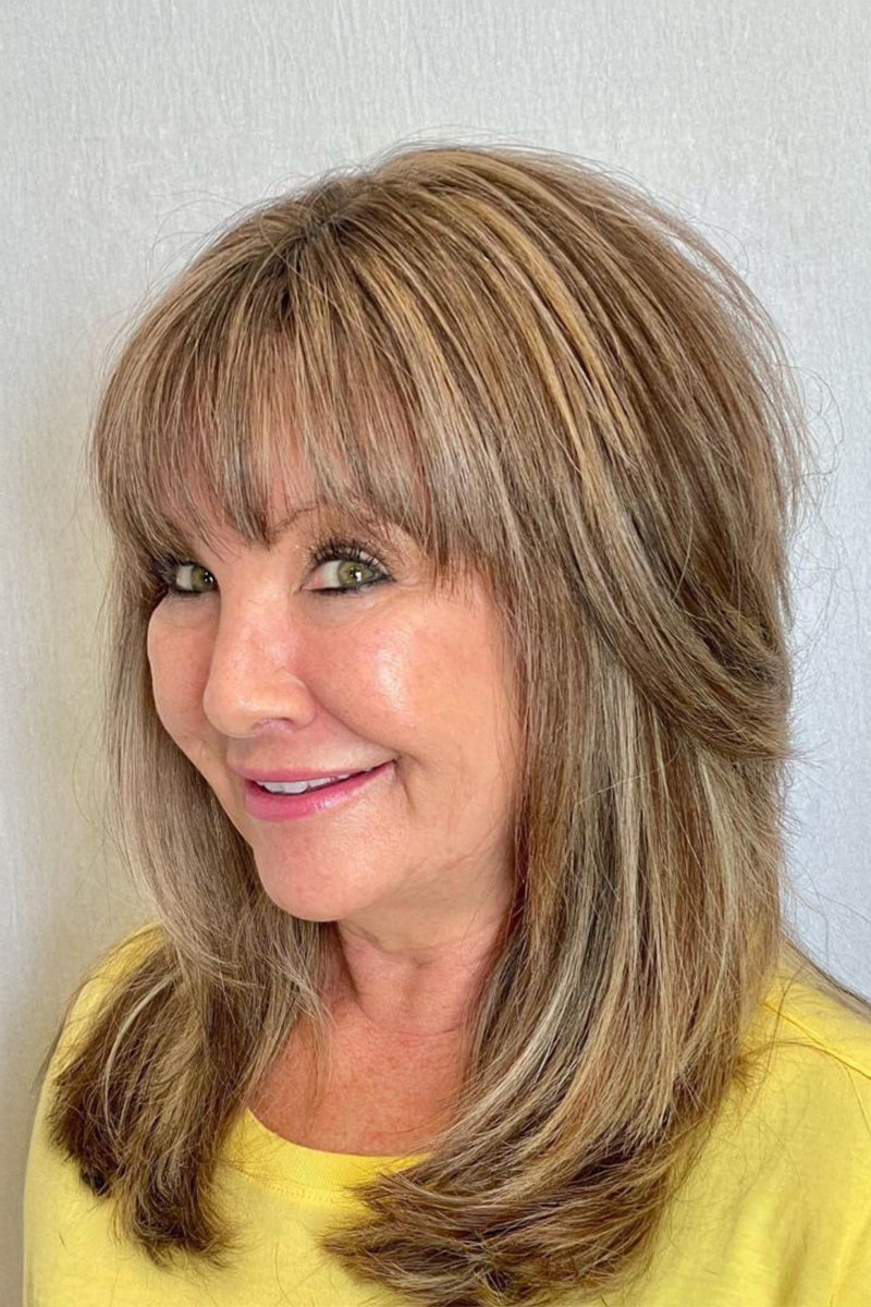 A close-up shot of a woman with shoulder-length layered hair featuring blonde highlights and straight-cut bangs, wearing a yellow top against a light gray wall.