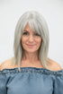 A woman with shoulder-length silver-gray hair and straight bangs, wearing an off-the-shoulder blue top against a plain light gray background.