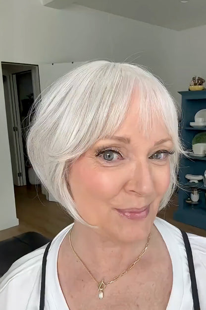 A woman with a short, layered bob wig in platinum white with soft gray lowlights, styled with straight-cut bangs, shown in a home setting.