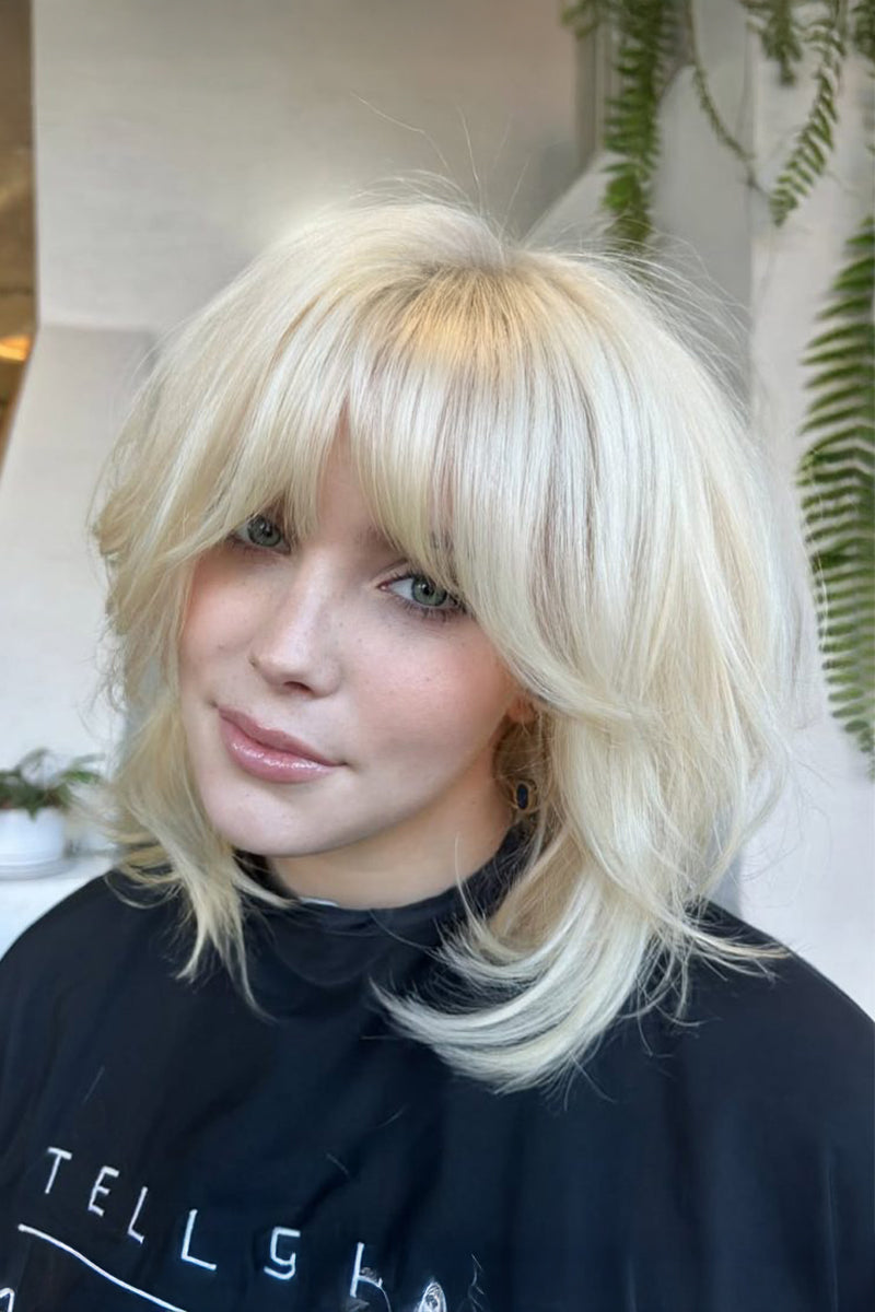 A close-up of a woman with a layered, shoulder-length platinum blonde wig with straight-cut bangs. She is wearing a black salon cape with white lettering, and the background shows a softly blurred indoor setting with green plants.