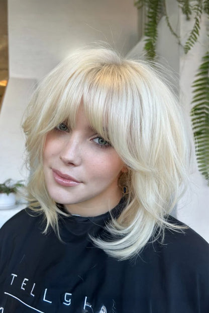 A close-up of a woman with a layered, shoulder-length platinum blonde wig with straight-cut bangs. She is wearing a black salon cape with white lettering, and the background shows a softly blurred indoor setting with green plants.